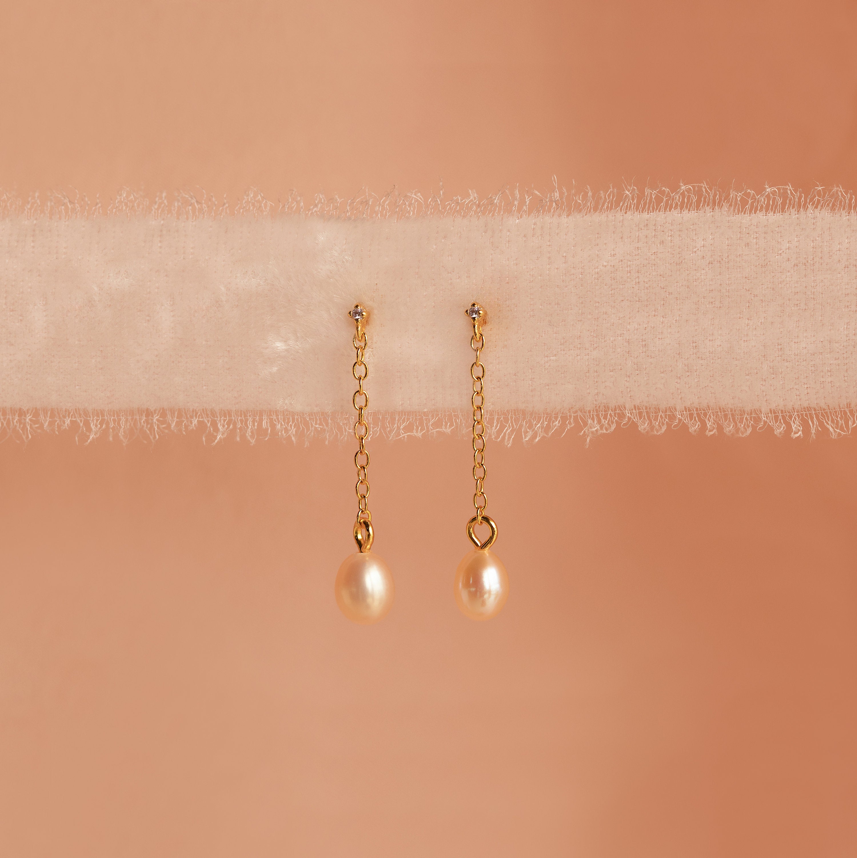 Isha Pink Pearl Earrings feature gold chains with pearl drops, elegantly displayed on soft, sheer fabric against a peach background—showcasing the timeless charm of these delicate dangling earrings.