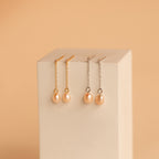 Showcased on a beige block against a peach background are two pairs of Isha Pink Pearl Earrings—one gold, one silver. One pair features delicate dangling chains for added elegance.