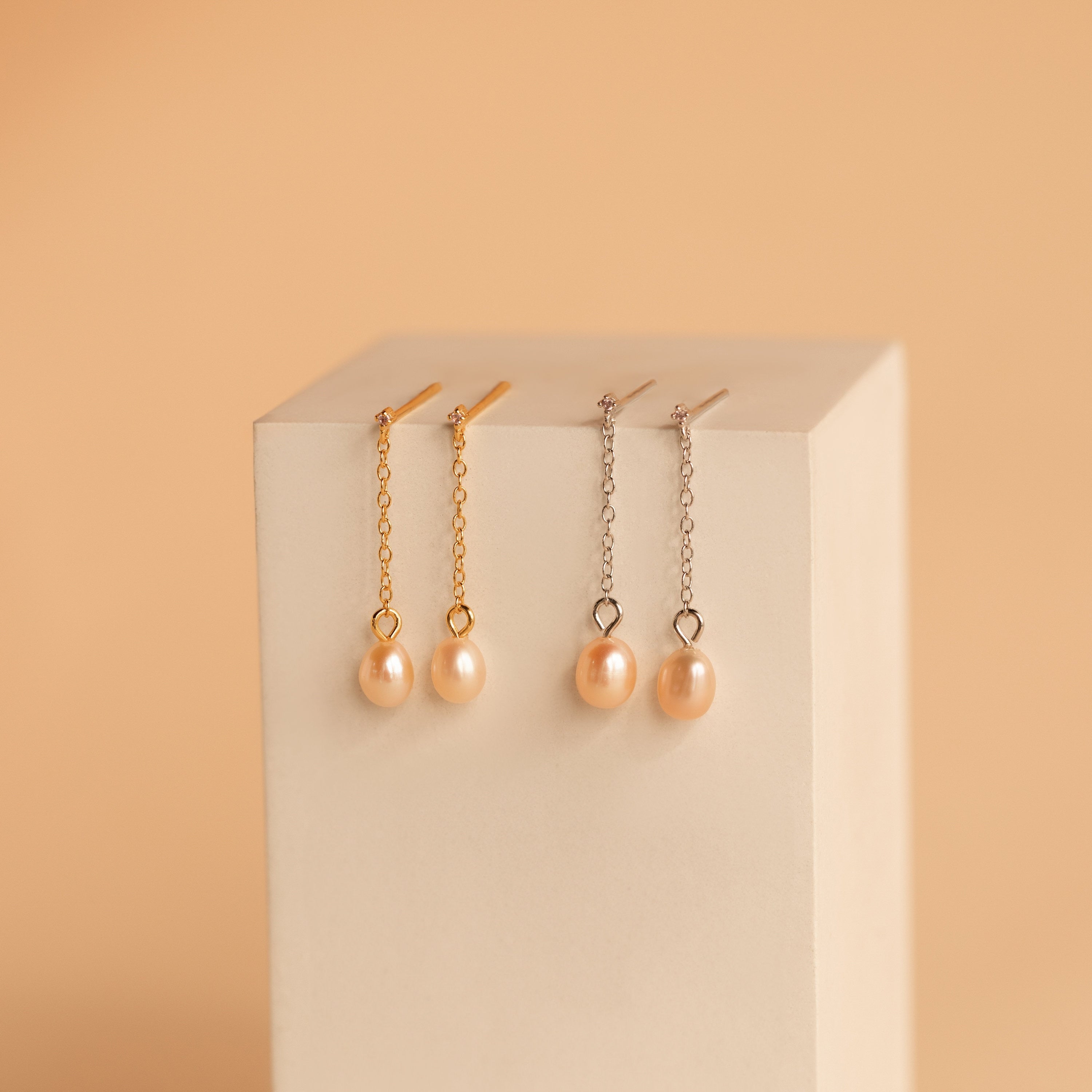 Showcased on a beige block against a peach background are two pairs of Isha Pink Pearl Earrings—one gold, one silver. One pair features delicate dangling chains for added elegance.