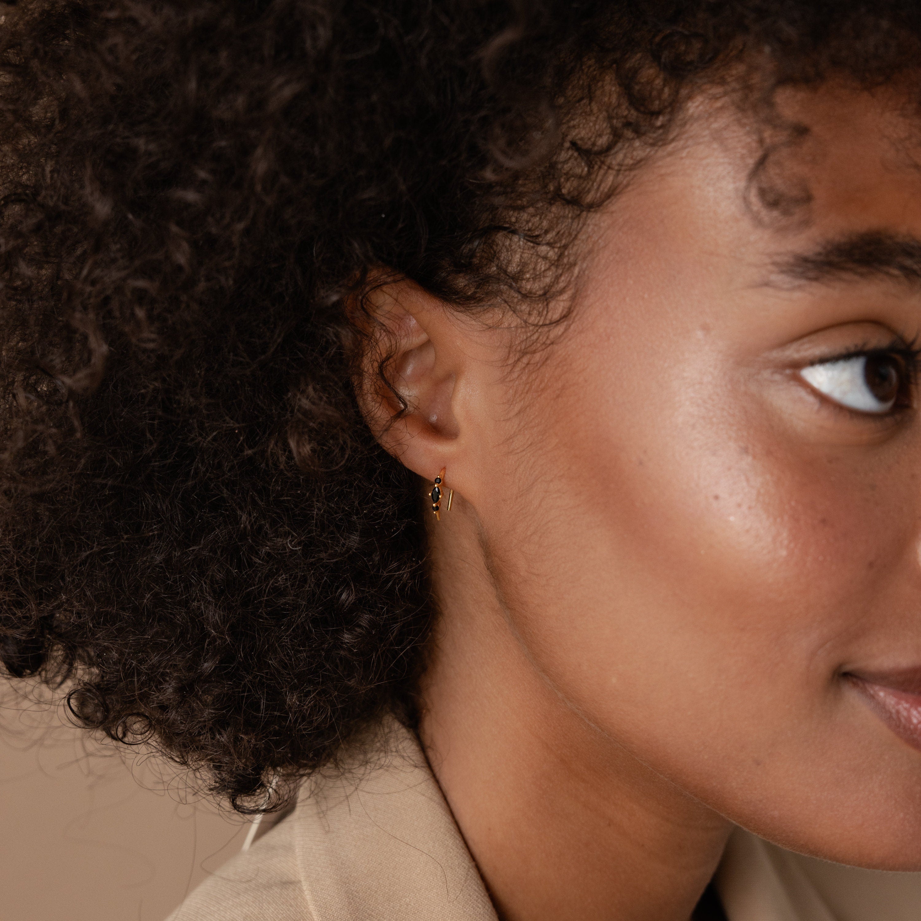 Close-up of a woman's face, highlighting her curly hair, smooth skin, and a gold hoop from our Amaya Black Marquise Threaders.