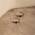 The Amaya Black Marquise Threaders, modern silver hoop earrings with black stones, are displayed on a beige marble surface.