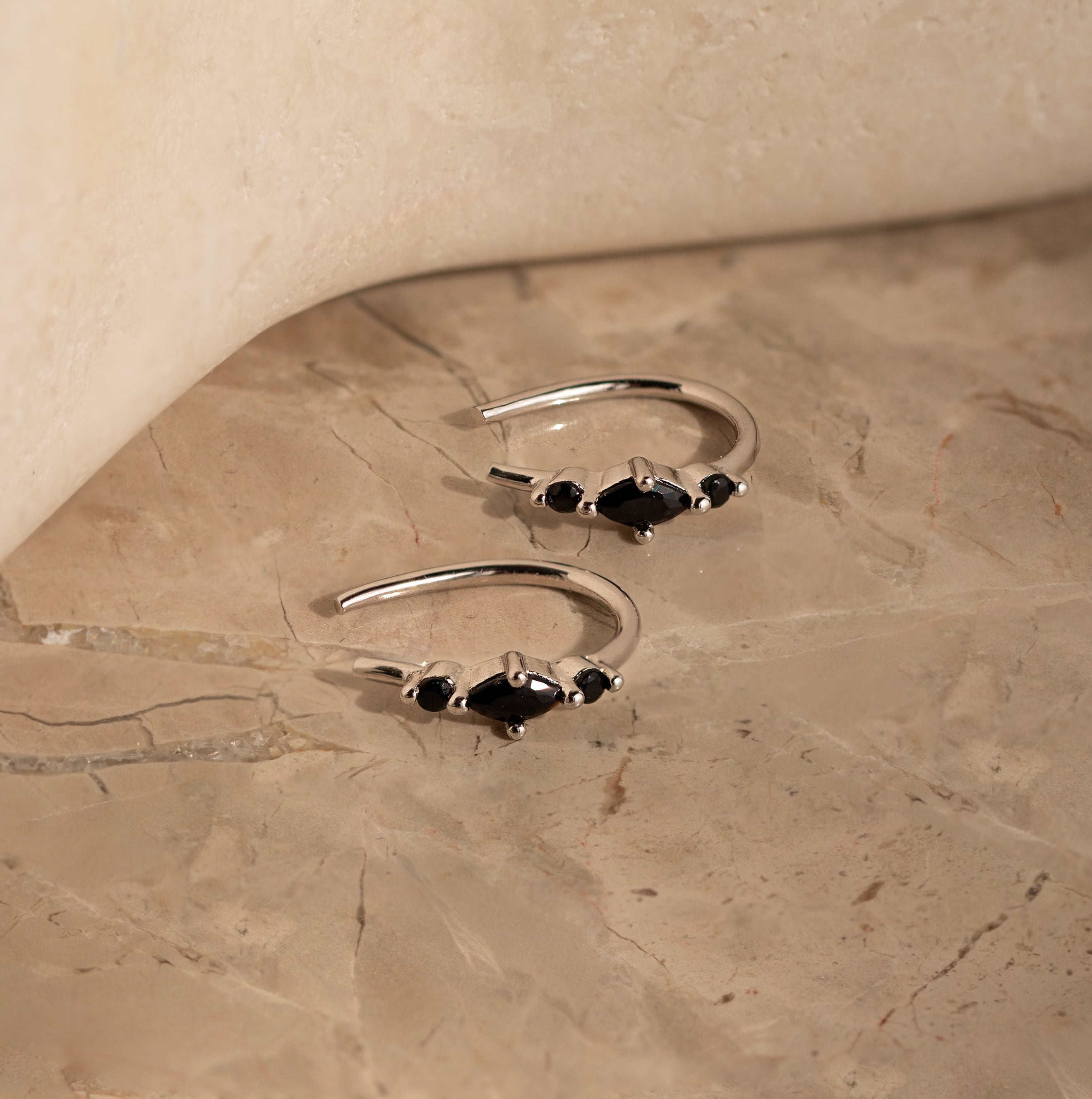 The Amaya Black Marquise Threaders, modern silver hoop earrings with black stones, are displayed on a beige marble surface.