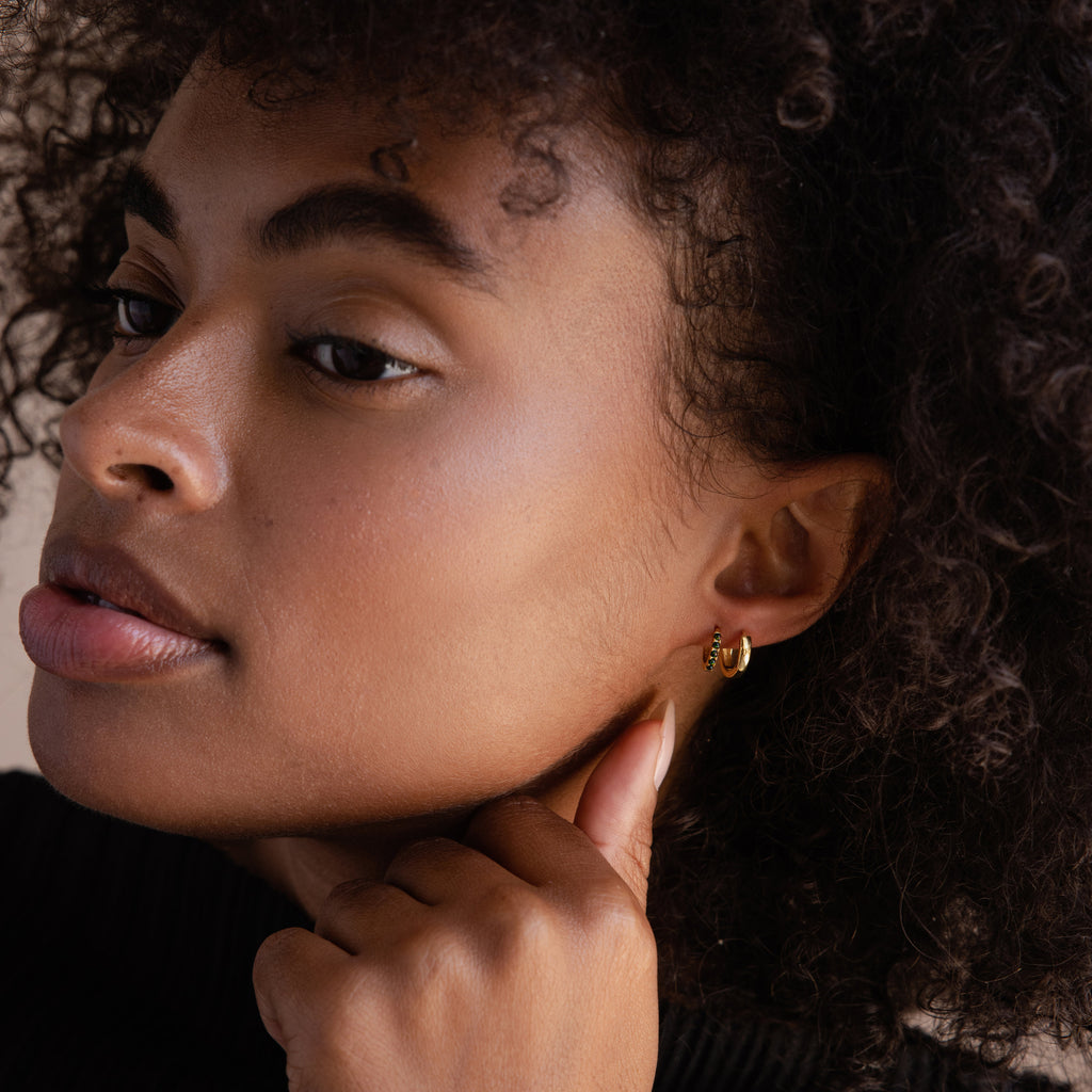 A woman with curly hair touches her ear and gazes thoughtfully to the side, showcasing the Black Sand Opal Huggies.