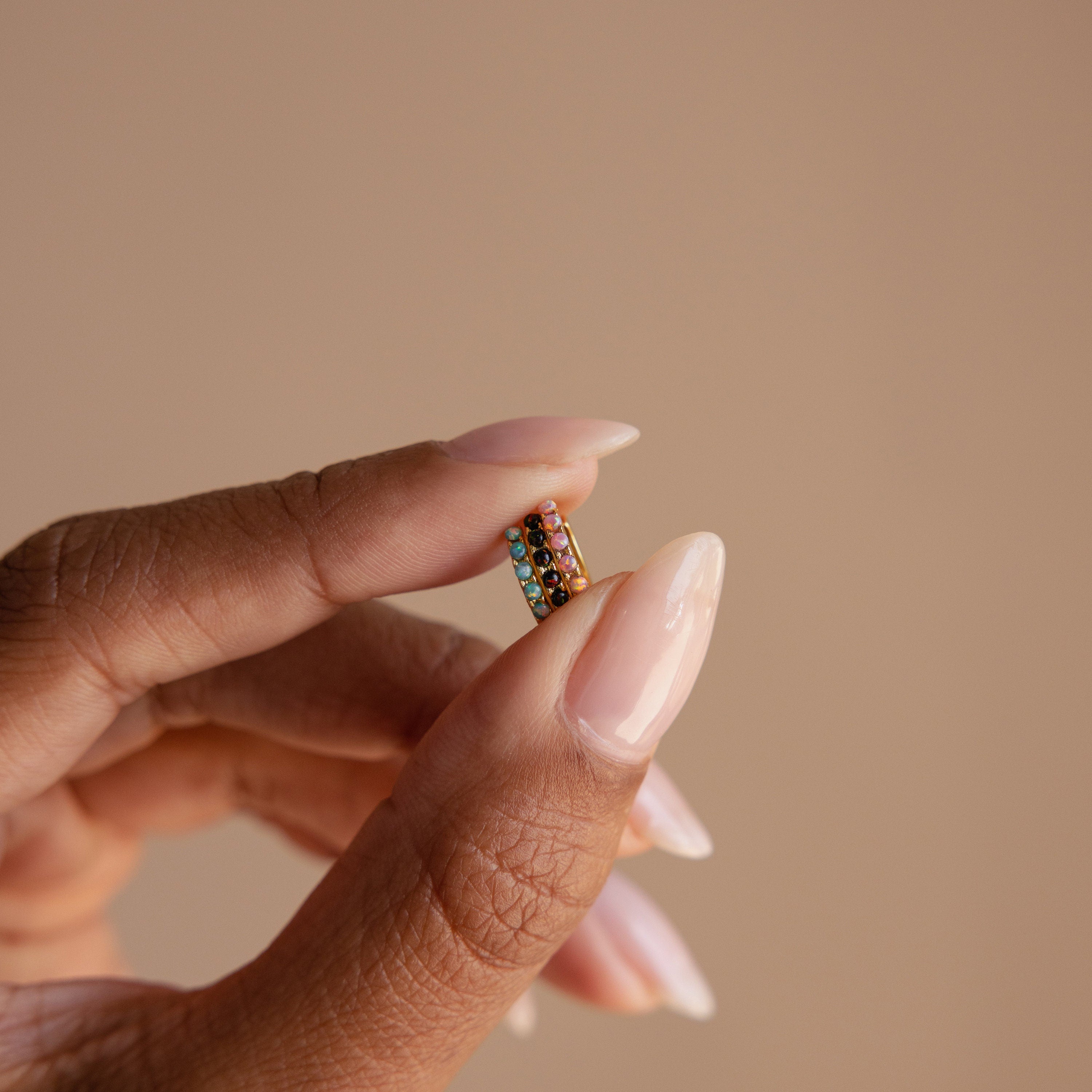 A hand with manicured nails displays the Black Sand Opal Huggies—small, colorful hoop earrings—against a neutral background.