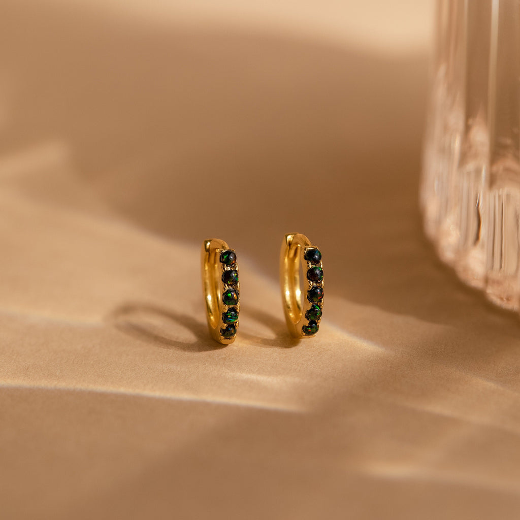 The Black Sand Opal Huggies—gold dainty hoop earrings with dark green and black stones—rest on a beige surface near a textured glass object, offering an elegant touch for everyday wear.