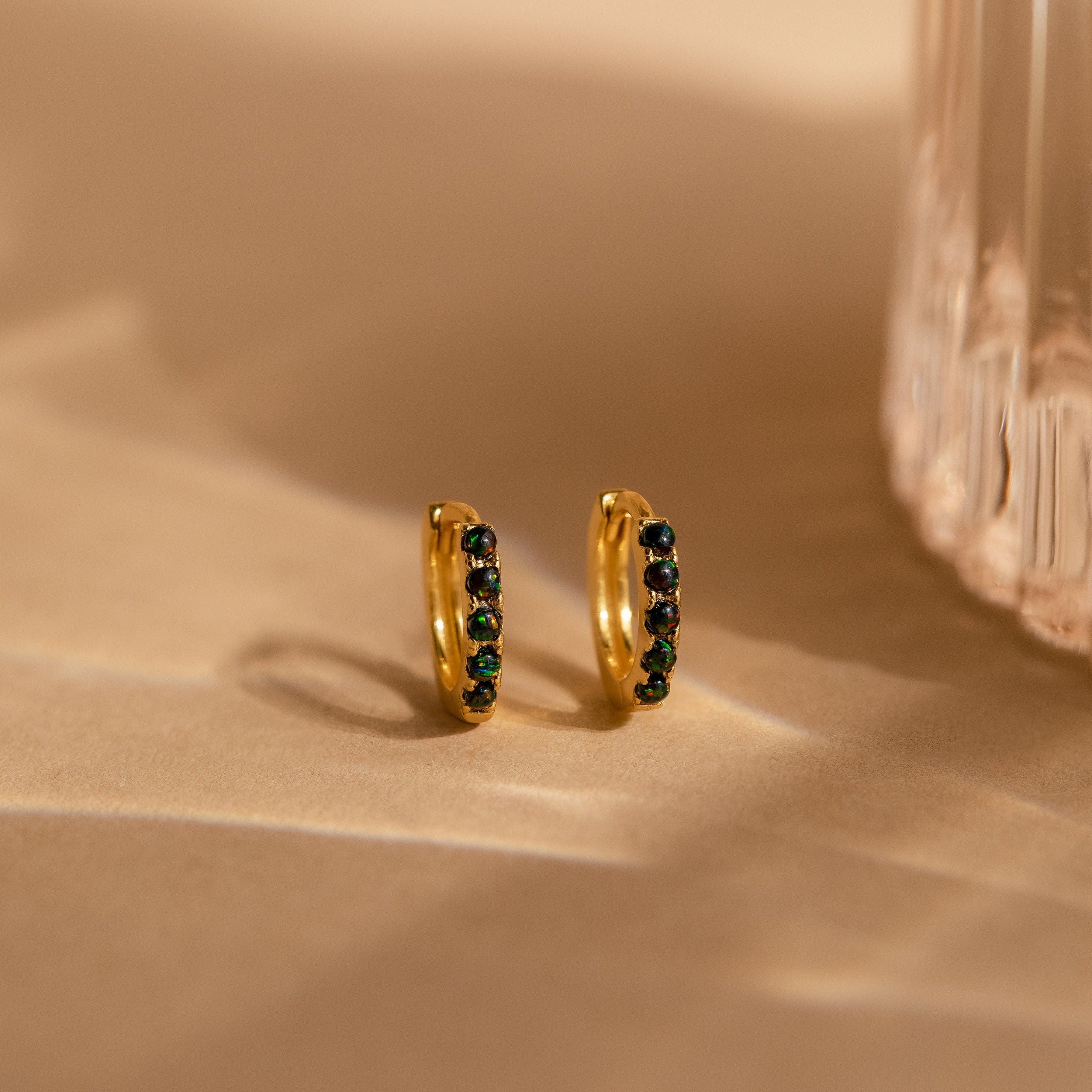 The Black Sand Opal Huggies—gold dainty hoop earrings with dark green and black stones—rest on a beige surface near a textured glass object, offering an elegant touch for everyday wear.