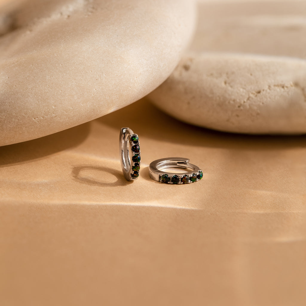The Black Sand Opal Huggies—dainty hoop earrings with green stones—rest on a beige surface beside smooth, rounded stones, making them the perfect everyday earrings for a touch of elegance.