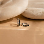 The Black Sand Opal Huggies—dainty hoop earrings with green stones—rest on a beige surface beside smooth, rounded stones, making them the perfect everyday earrings for a touch of elegance.