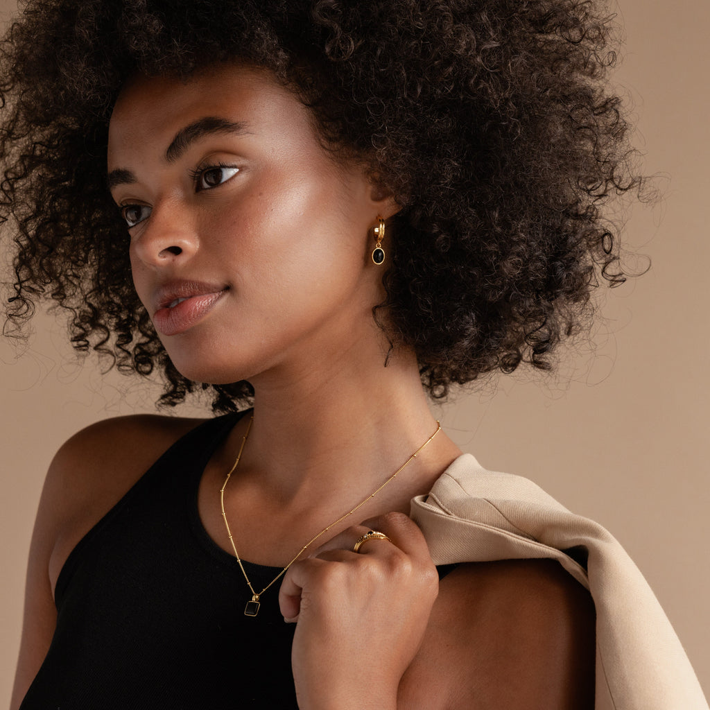 Woman with curly hair wearing Black Gemstone Drop Hoops, a black top, and a beige jacket draped over her shoulder.