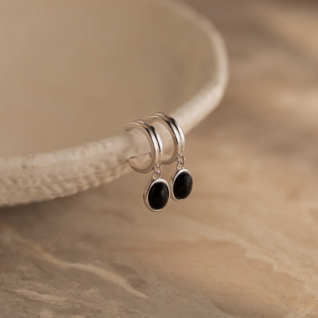 Black Gemstone Drop Hoops with dangling black oval stones rest on a textured beige dish—perfect as stylish everyday earrings or striking black gemstone accents for any occasion.