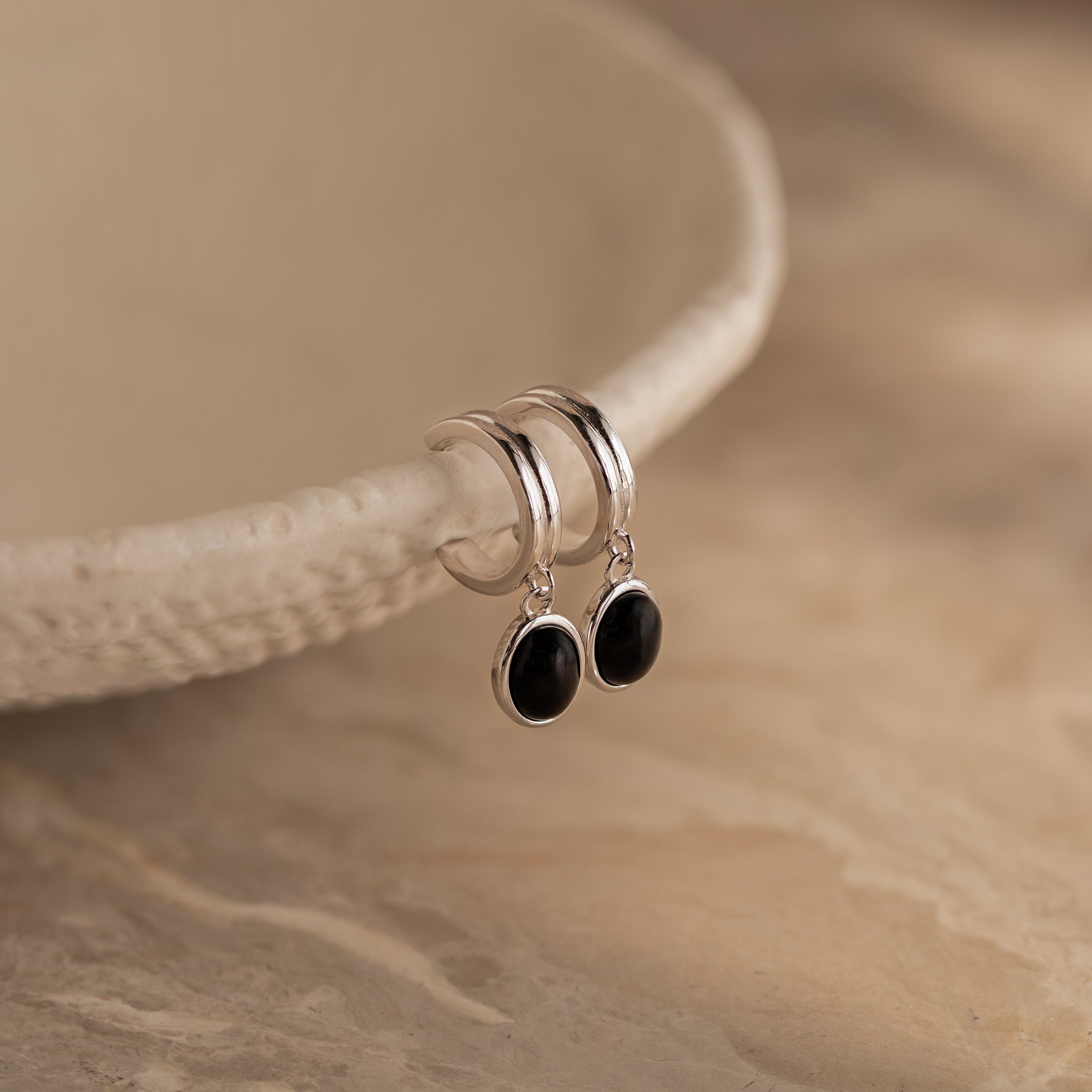 Black Gemstone Drop Hoops with dangling black oval stones rest on a textured beige dish—perfect as stylish everyday earrings or striking black gemstone accents for any occasion.