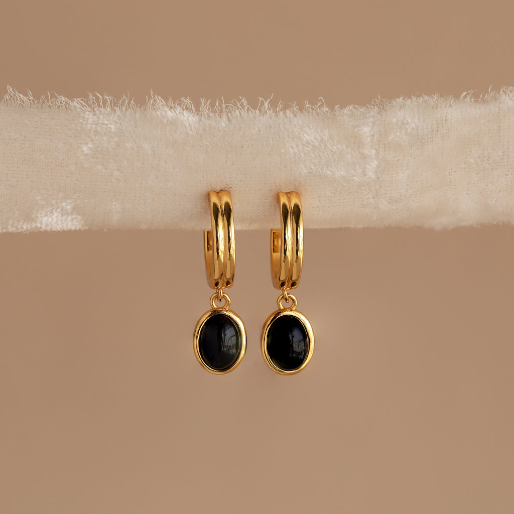Black Gemstone Drop Hoops featuring gold hoops with black round gemstone pendants, shown on a soft beige fabric background. Ideal everyday earrings for a subtle, elegant look.