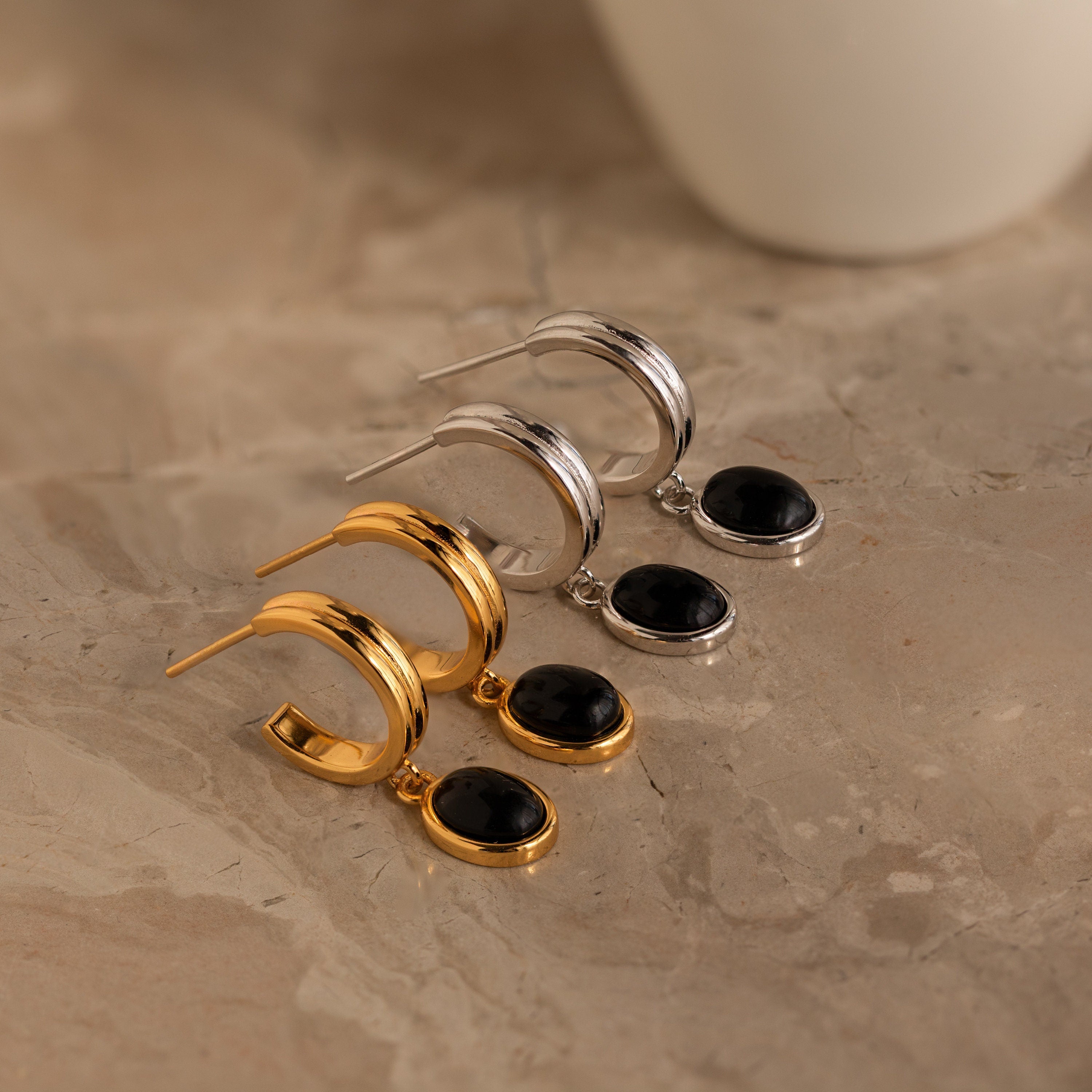 Two pairs of Black Gemstone Drop Hoops, one gold and one silver, each with a black gemstone pendant, on a marble surface. Ideal for adding everyday elegance to any look.