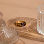 The Dark Amethyst Signet Ring, featuring a striking purple gemstone set in gold, is displayed on a beige surface and accented by decorative glass pieces casting elegant shadows.