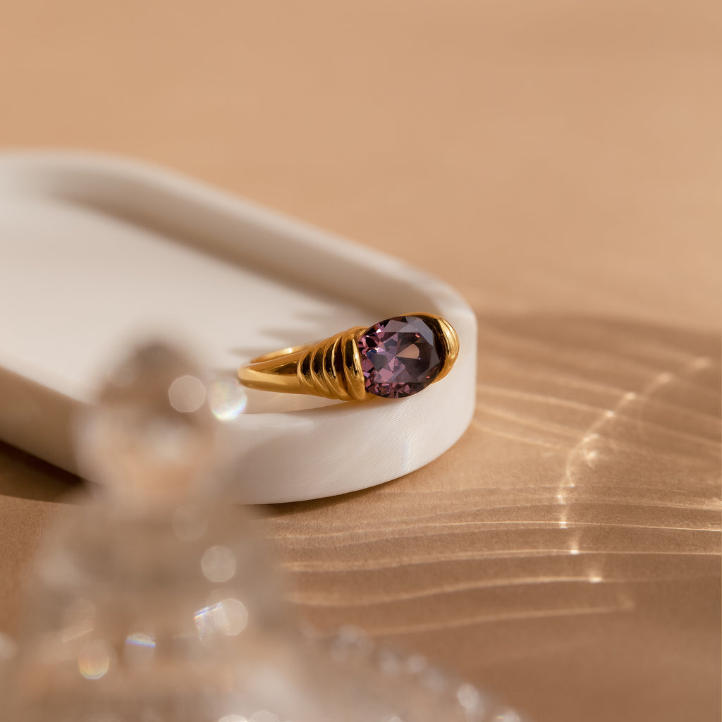 The Dark Amethyst Signet Ring showcases a vibrant purple gemstone, displayed on a white tray with soft shadows over a beige surface—a vintage-inspired piece that exudes timeless elegance.