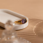 The Dark Amethyst Signet Ring showcases a vibrant purple gemstone, displayed on a white tray with soft shadows over a beige surface—a vintage-inspired piece that exudes timeless elegance.