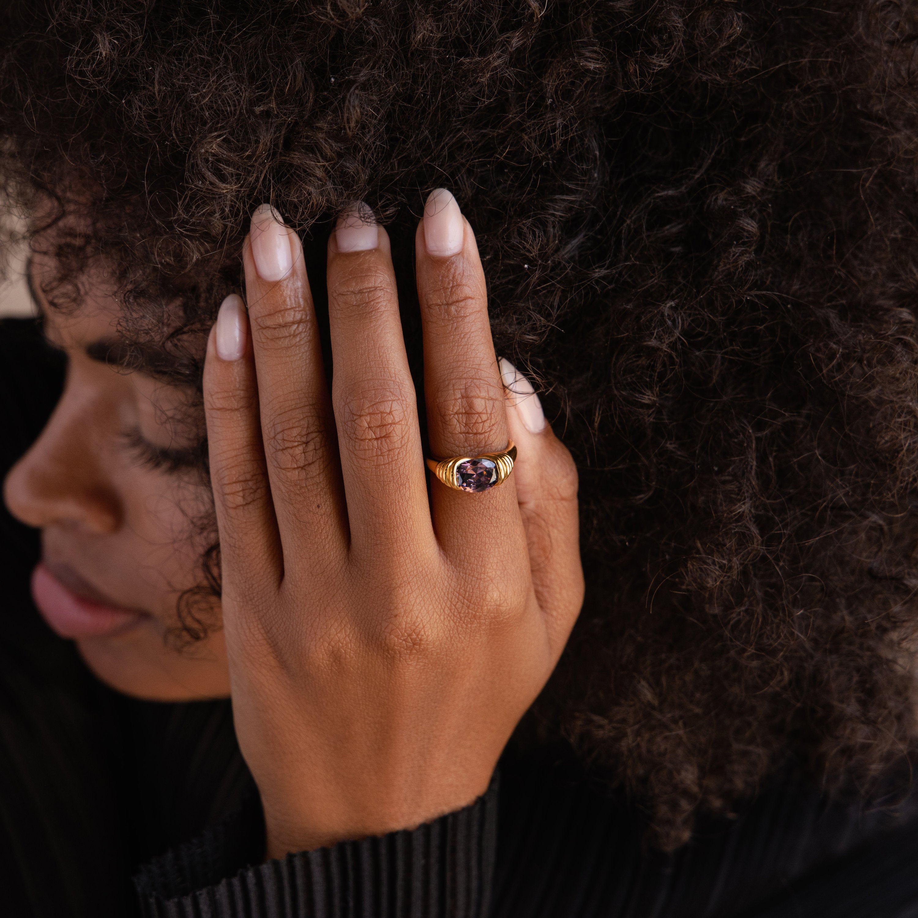A person with curly hair touches their face, revealing a gold Dark Amethyst Signet Ring on their finger.