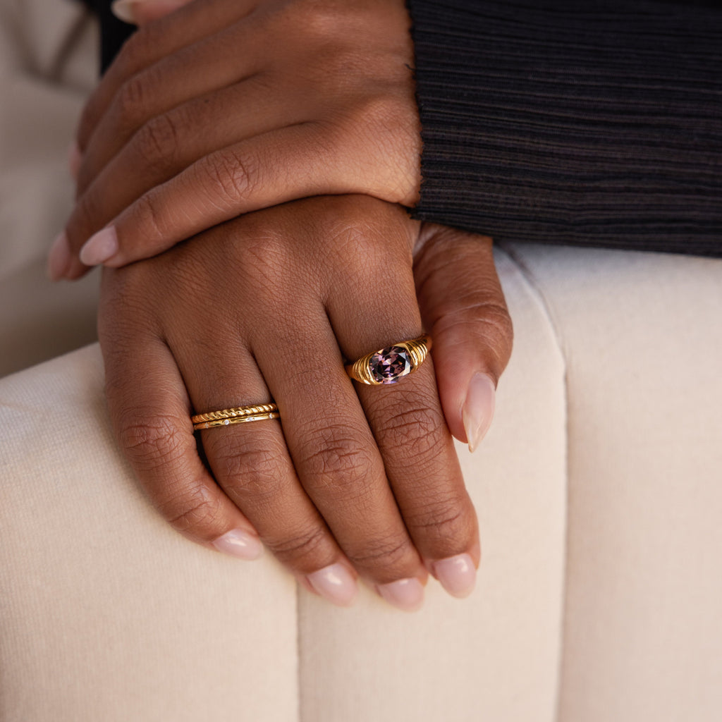 Two hands rest on a chair; one showcases manicured nails and two gold rings, including the Dark Amethyst Signet Ring, a vintage-inspired piece featuring a striking purple stone.