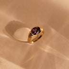The Dark Amethyst Signet Ring is a vintage-inspired gold band featuring a purple oval stone, reminiscent of Art Deco style and casting a soft shadow on a beige surface.