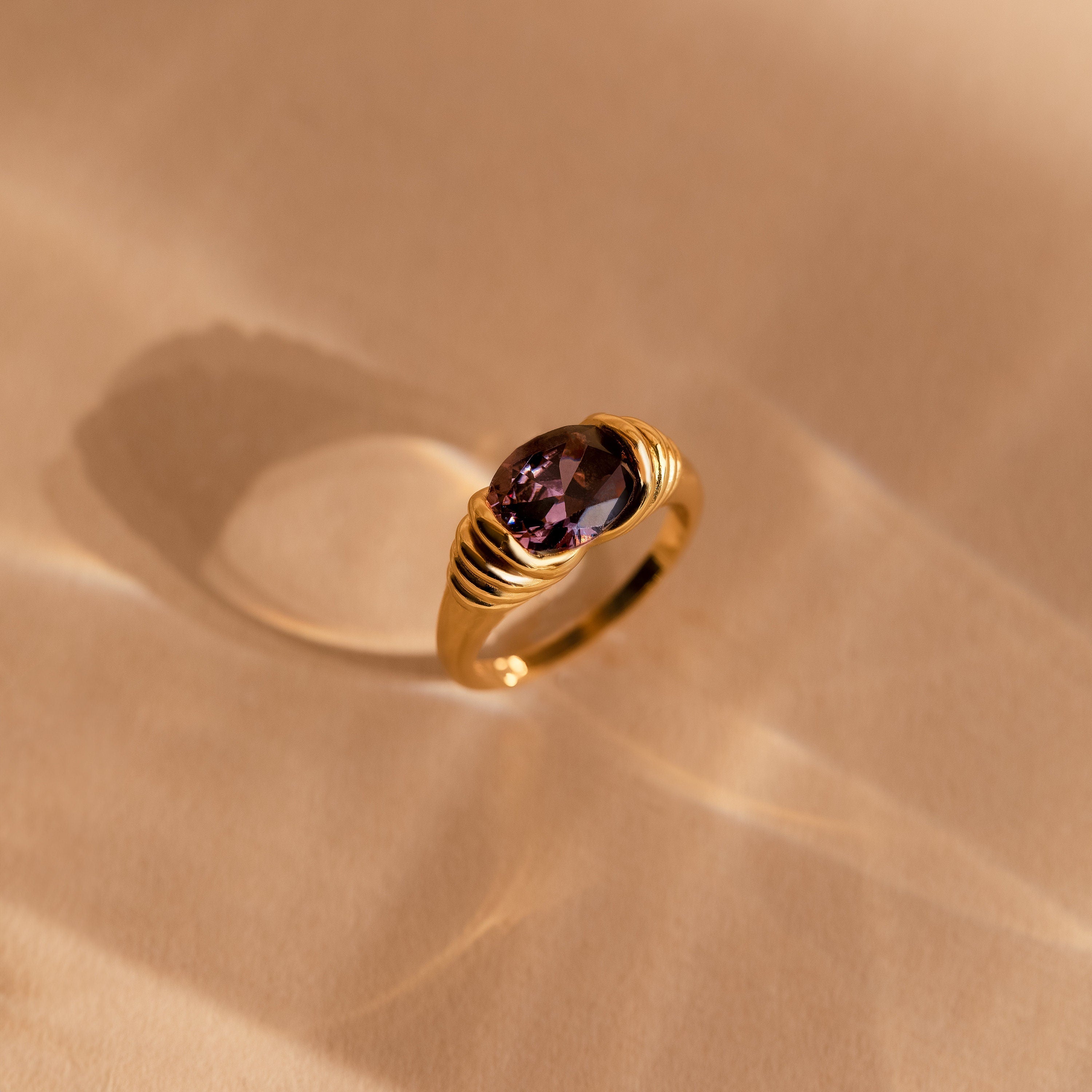 The Dark Amethyst Signet Ring is a vintage-inspired gold band featuring a purple oval stone, reminiscent of Art Deco style and casting a soft shadow on a beige surface.