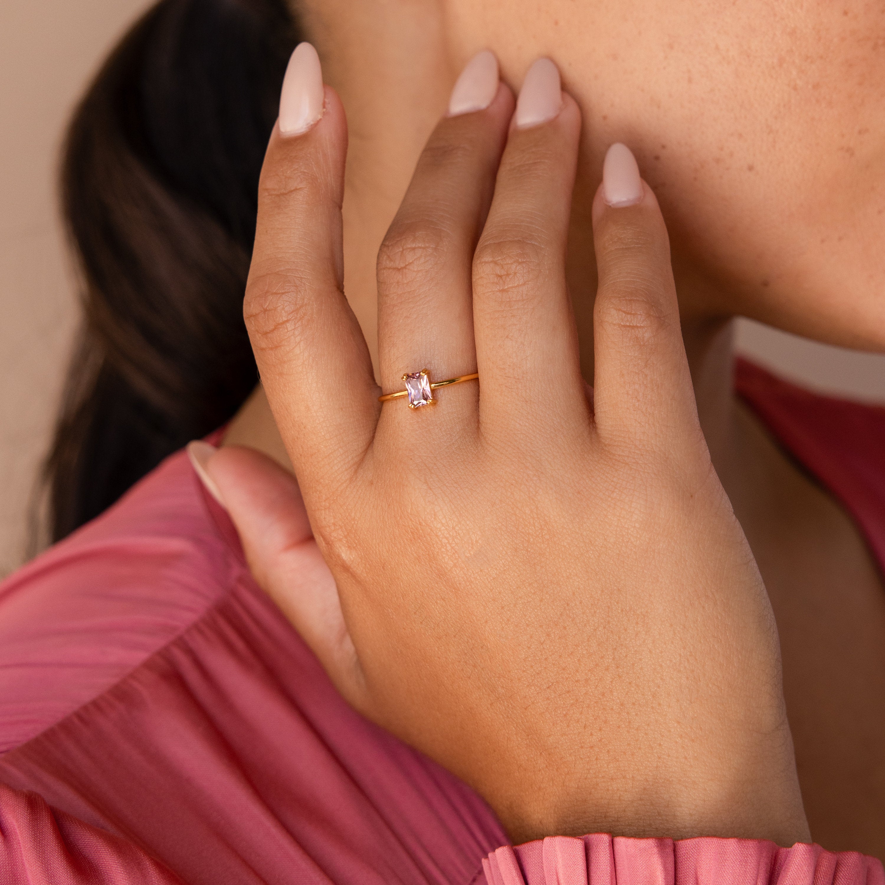 A hand with pale pink nails wears the Blush Pink Emerald Ring, a dainty stackable piece with a rectangular gemstone, near a face in a pink top.