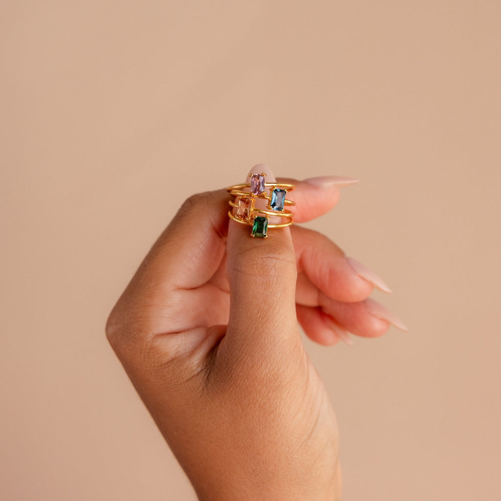 A hand with long nails showcases three stackable rings, including the Blush Pink Emerald Ring, each featuring a unique gemstone color.