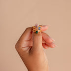 A hand with long nails showcases three stackable rings, including the Blush Pink Emerald Ring, each featuring a unique gemstone color.