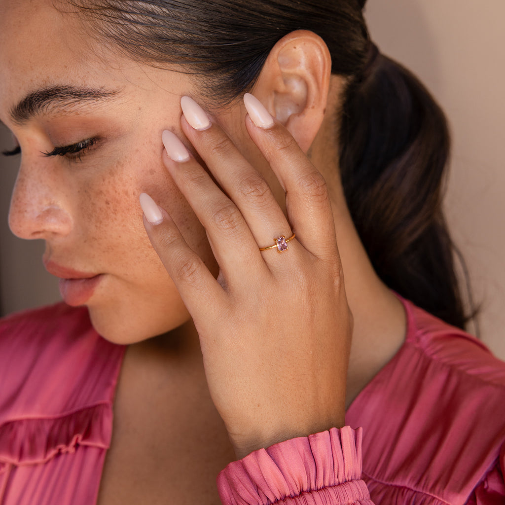 A woman with a ponytail and pink nails displays the Blush Pink Emerald Ring, featuring a delicate pink stone, while wearing a matching pink top.