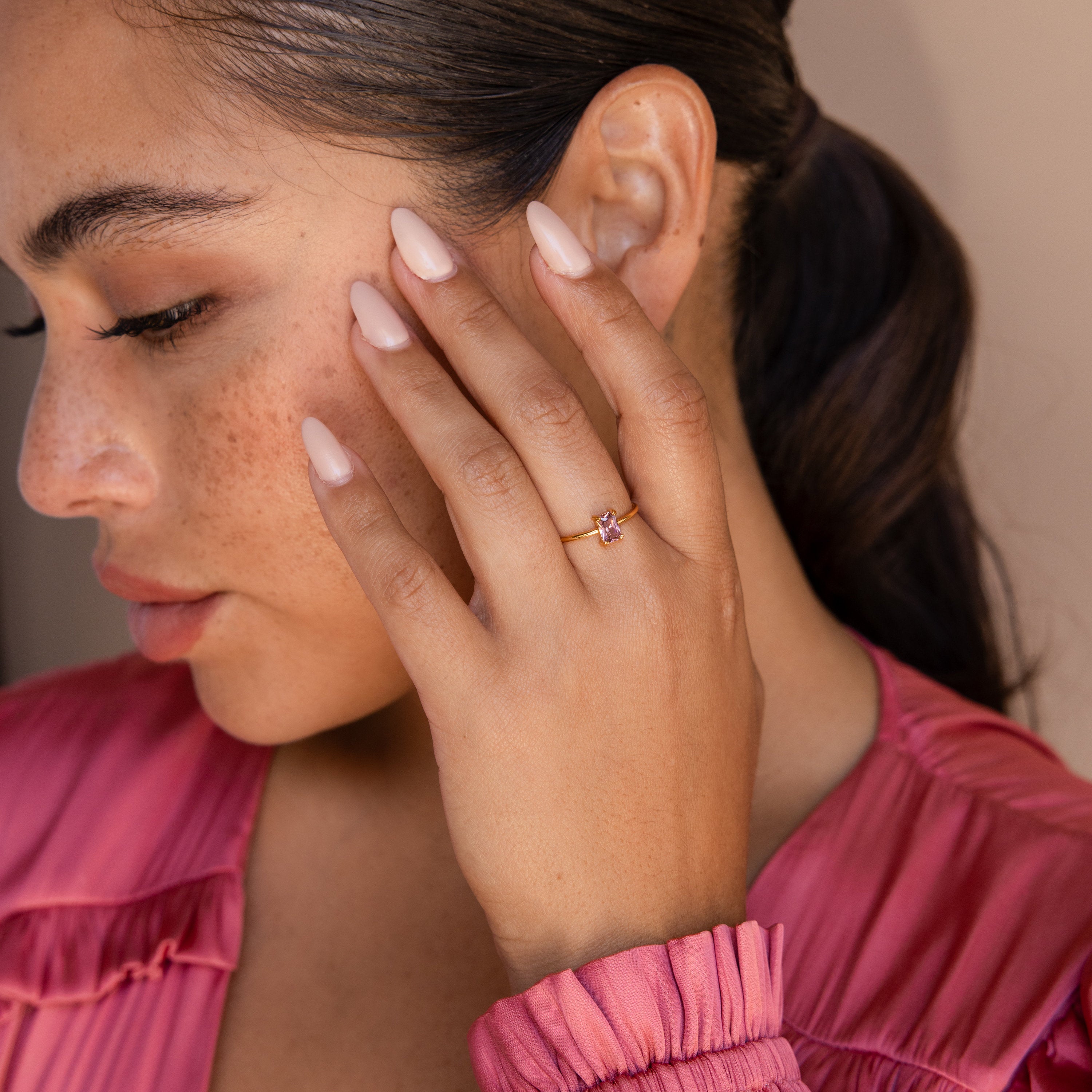 A woman with a ponytail and pink nails displays the Blush Pink Emerald Ring, featuring a delicate pink stone, while wearing a matching pink top.
