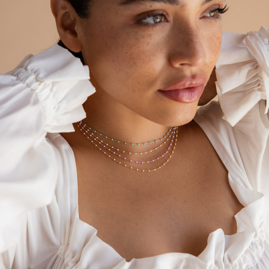 Woman in a white ruffled top wears layered, colorful bead necklaces—featuring the Pink Station Necklace—as she gazes off to the side.