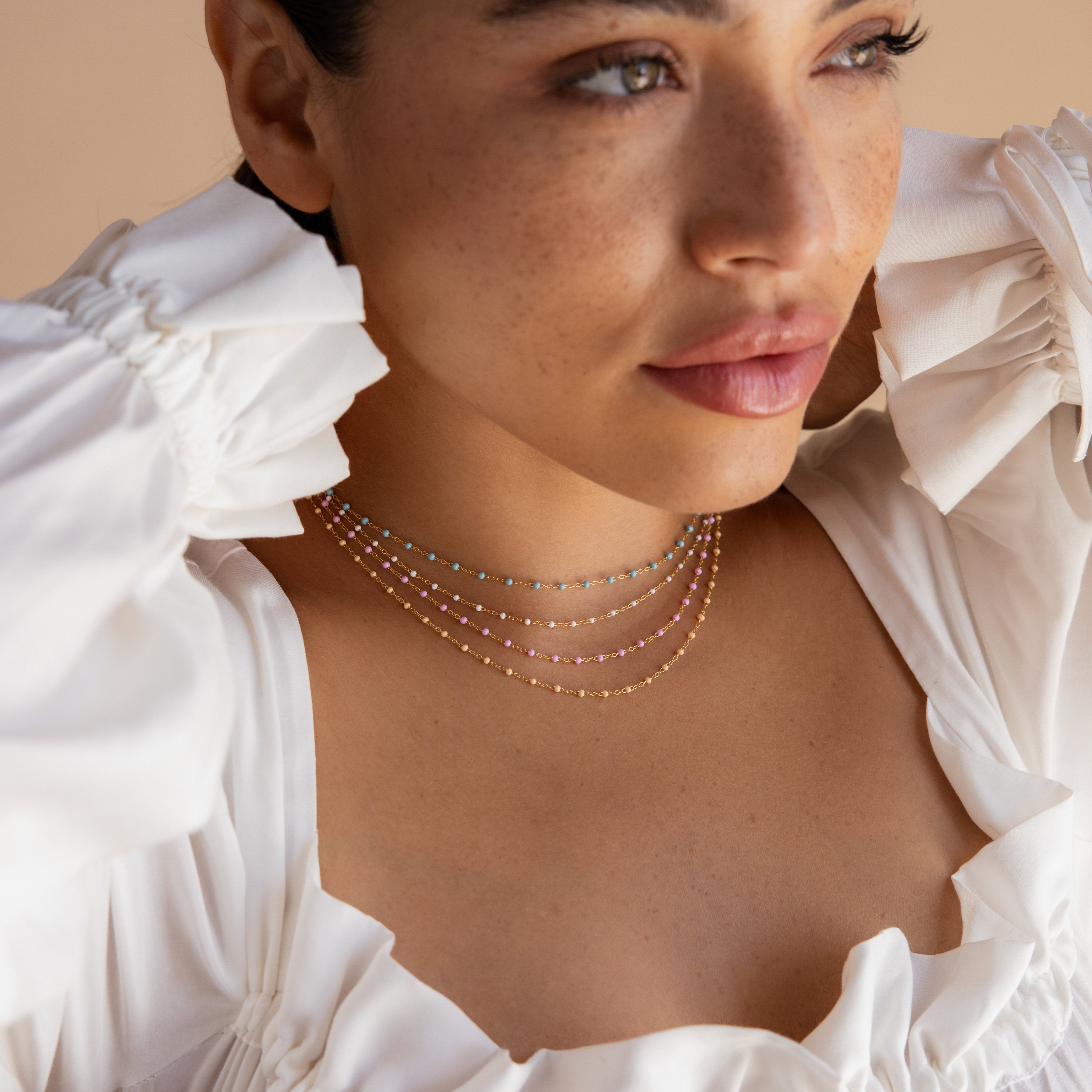 Woman in a white ruffled top wears layered, colorful bead necklaces—featuring the Pink Station Necklace—as she gazes off to the side.