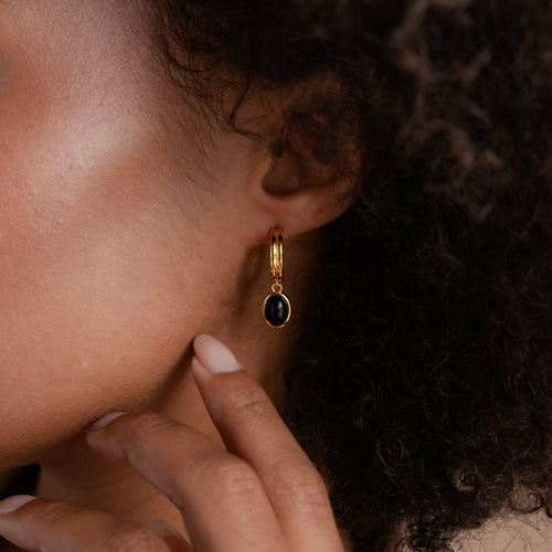Close-up of a person with curly hair wearing elegant Black Gemstone Drop Hoops in a gold oval setting, thoughtfully touching their chin. Perfect chic Everyday Earrings.