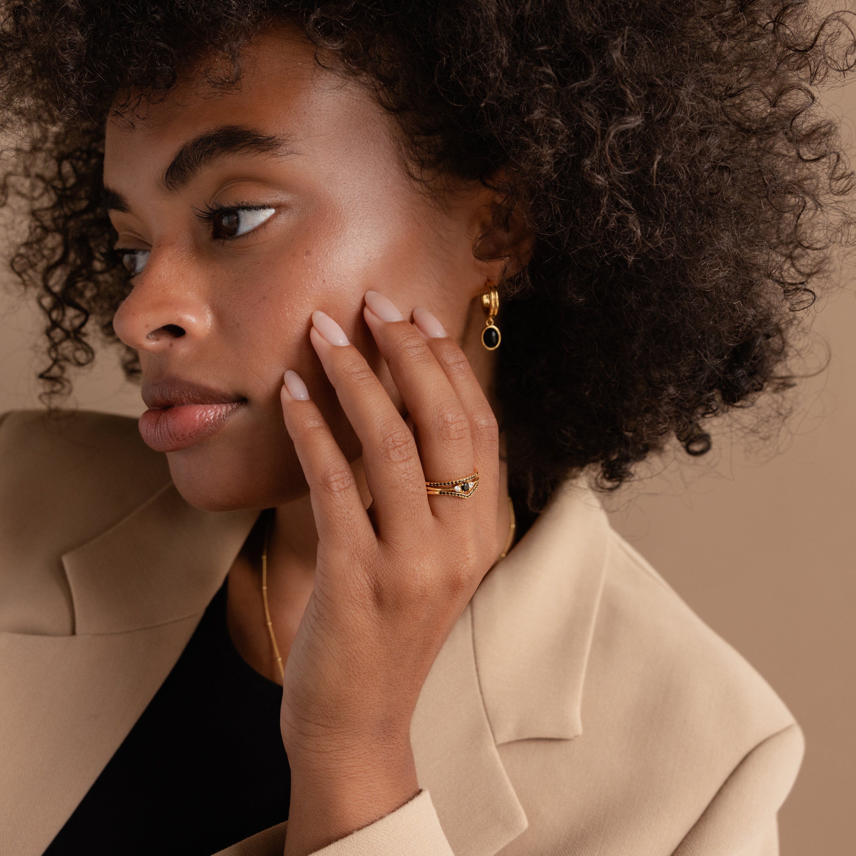 Woman with curly hair in a beige blazer and gold jewelry wears the elegant Black Curve Ring Set, touching her face with manicured nails.