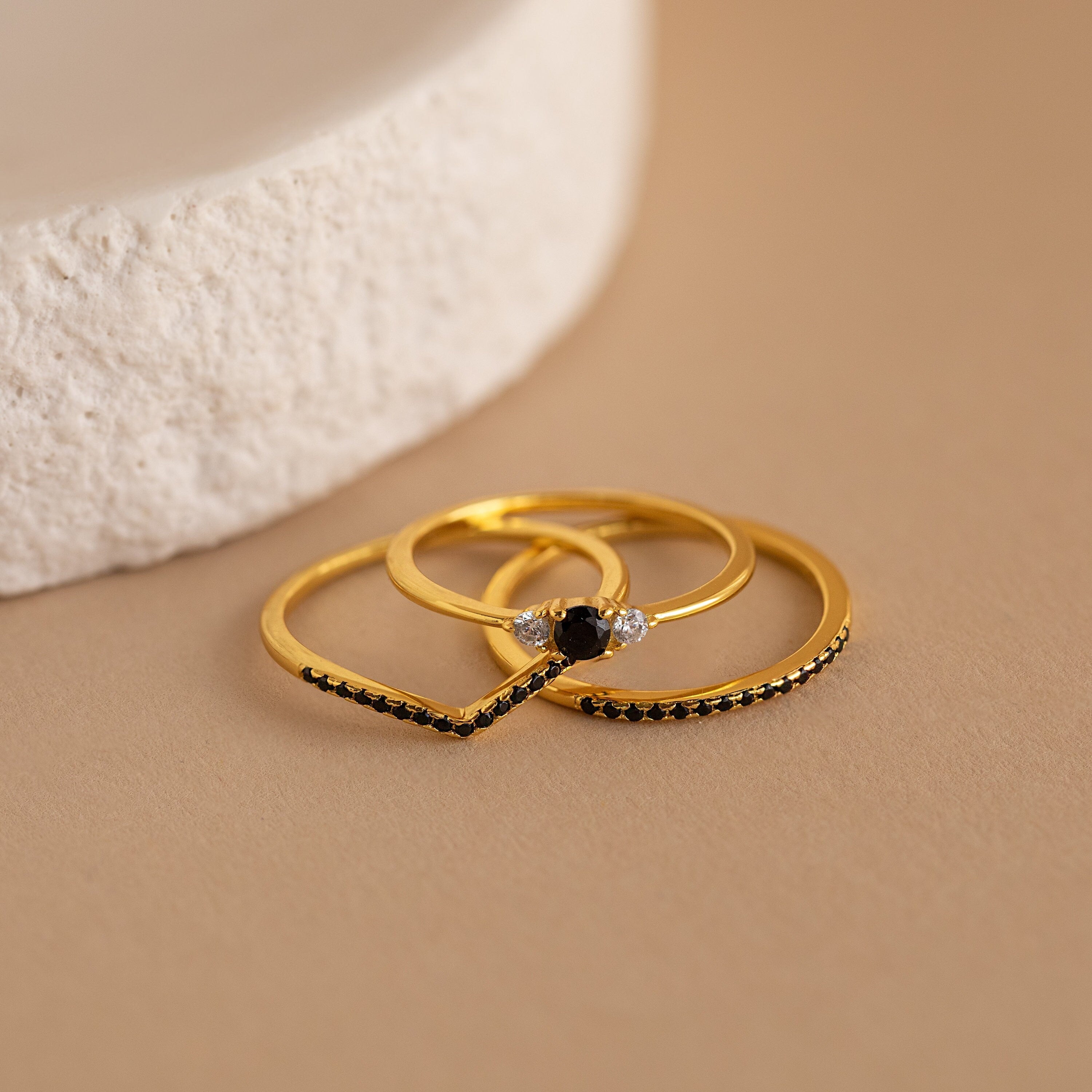 The Black Curve Ring Set, featuring three gold rings with black and diamond accents, is displayed on a beige surface beside a white textured item.