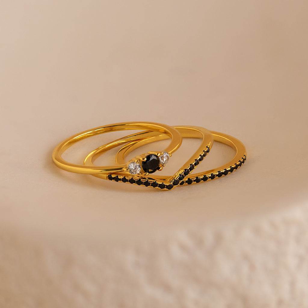 The Black Curve Ring Set features three elegant gold rings—a sparkling eternity band and two bold black diamond rings—stacked together on a subtly textured surface.