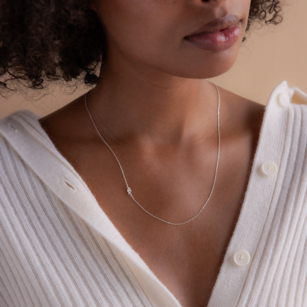 A woman in a cream sweater wears the Venice Sideways Initial Necklace, featuring a personalized silver initial charm for a delicate touch.