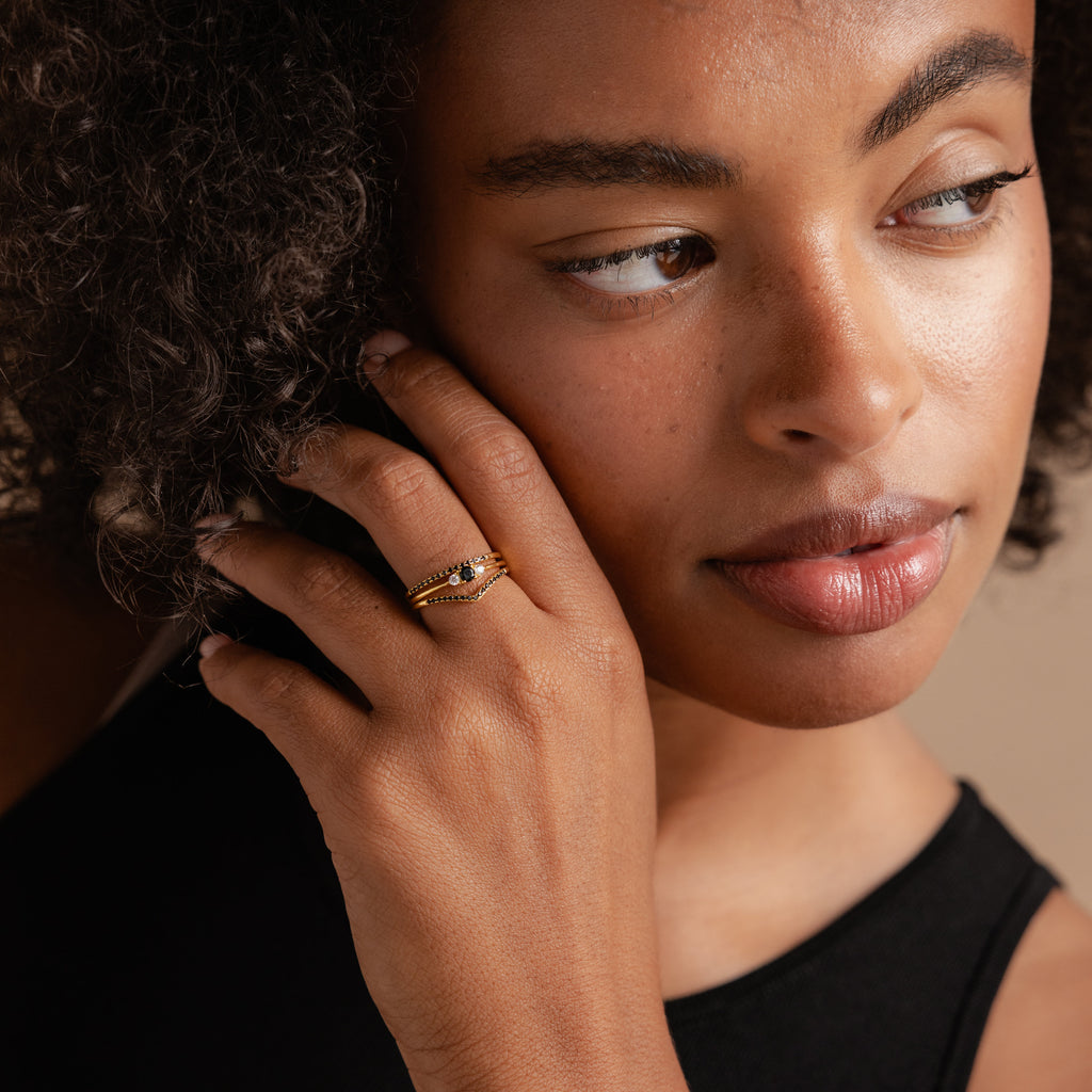 A woman with curly hair touches her face, highlighting the Black Curve Ring Set with dark stones, complemented by a sleeveless black top.