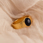 The Celestial Black Signet Ring features a textured gold band and a round black stone, creating a striking statement piece that stands out beautifully against soft beige fabric.