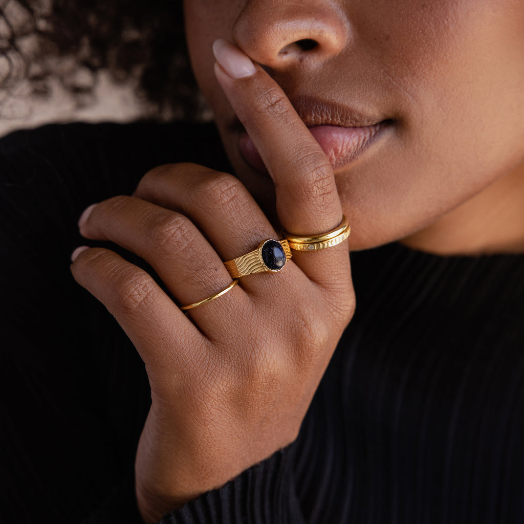 A woman wearing gold rings, including the Celestial Black Signet Ring, holds her finger to her lips in a gesture of silence—an elegant tribute to statement jewelry.