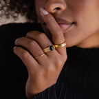 A woman wearing gold rings, including the Celestial Black Signet Ring, holds her finger to her lips in a gesture of silence—an elegant tribute to statement jewelry.