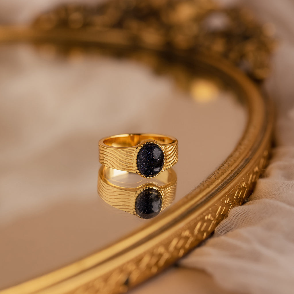The Celestial Black Signet Ring, featuring a dark oval stone, makes a striking statement and is elegantly displayed on an ornate gold-edged mirror.