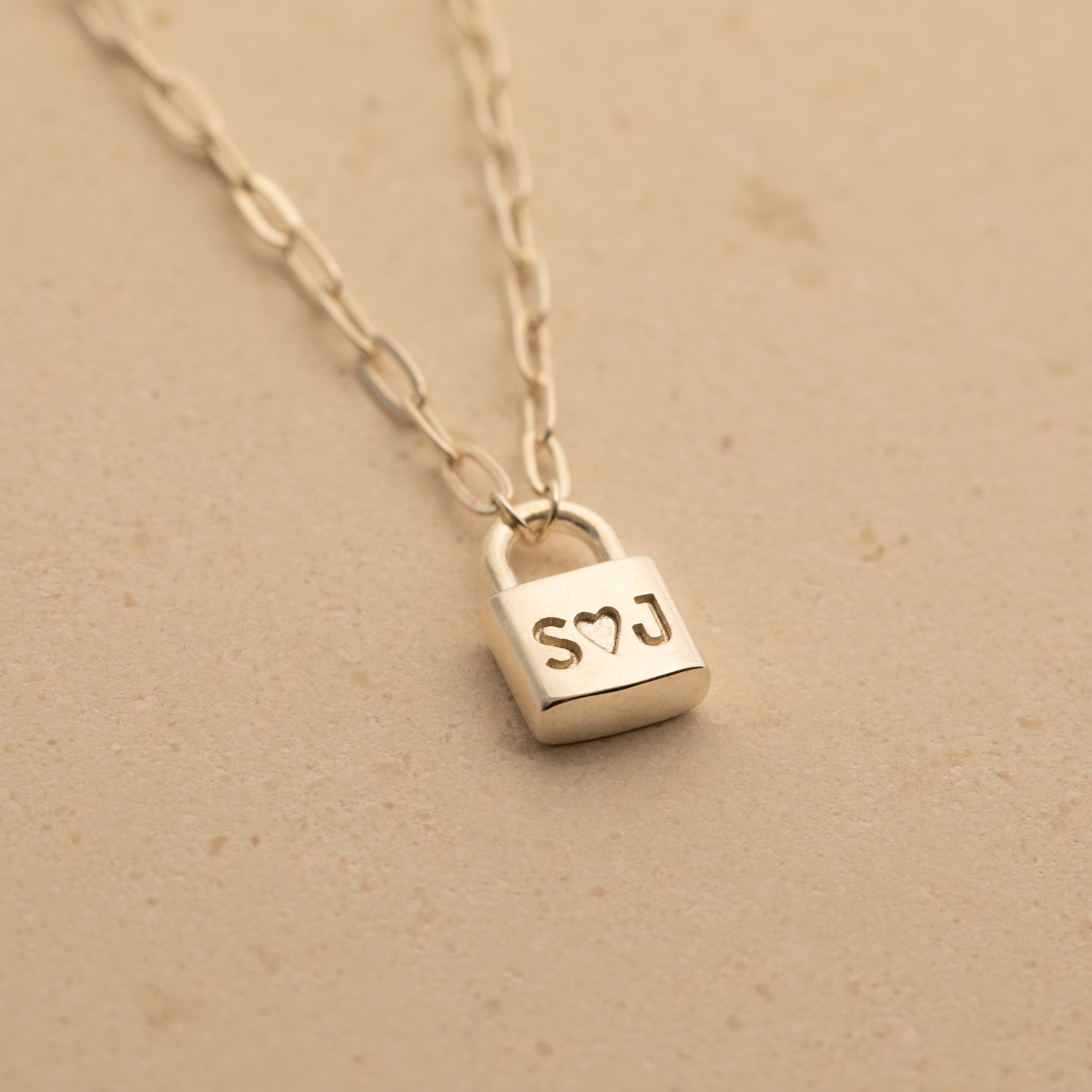 Initial Padlock Paperclip Necklace featuring a personalized padlock pendant engraved with S♡J, perfect for couples and beautifully displayed on a beige surface.