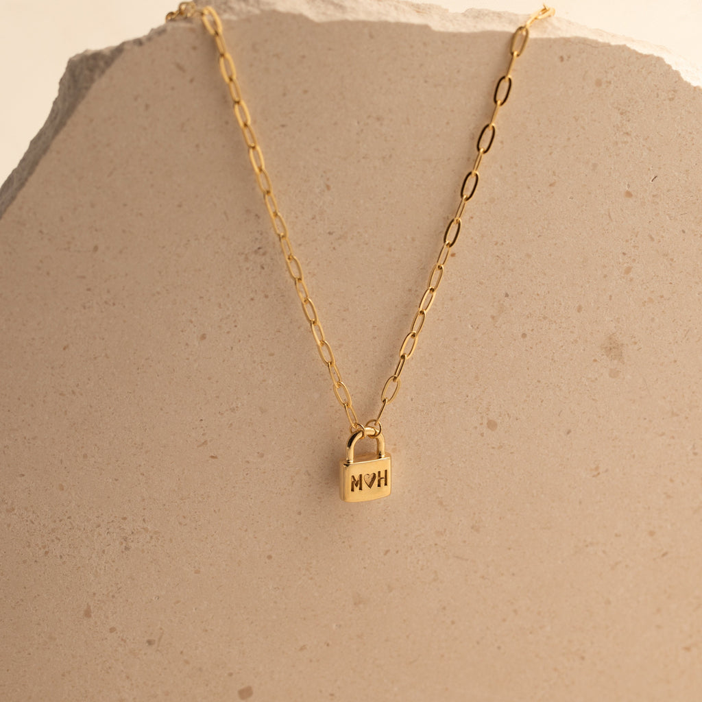 The Initial Padlock Paperclip Necklace features a gold chain with a small padlock pendant engraved with "MOM," displayed on a beige stone surface—a thoughtful personalized gift for any occasion.