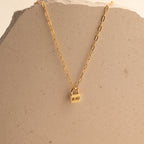 The Initial Padlock Paperclip Necklace features a gold chain with a small padlock pendant engraved with "MOM," displayed on a beige stone surface—a thoughtful personalized gift for any occasion.