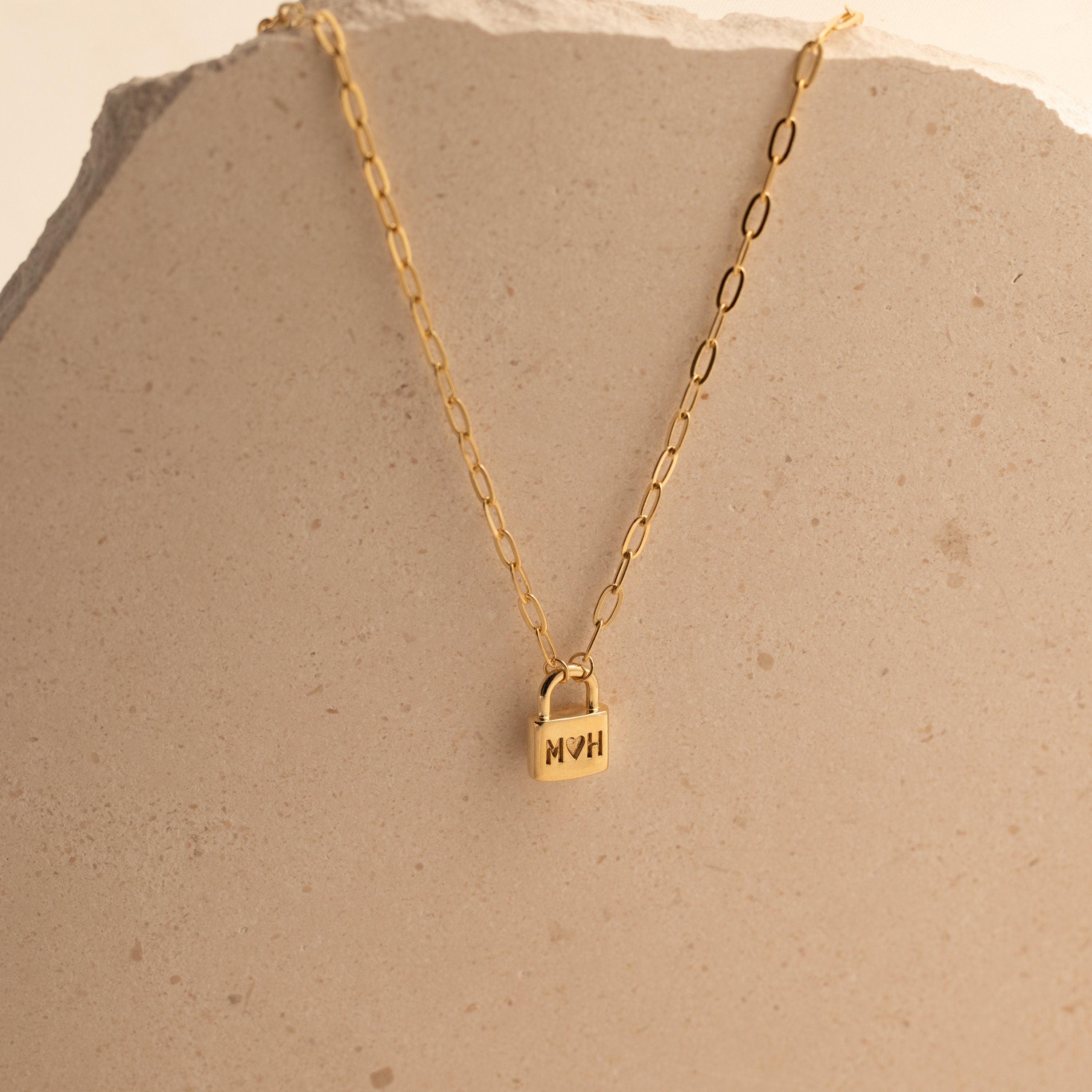 The Initial Padlock Paperclip Necklace features a gold chain with a small padlock pendant engraved with "MOM," displayed on a beige stone surface—a thoughtful personalized gift for any occasion.
