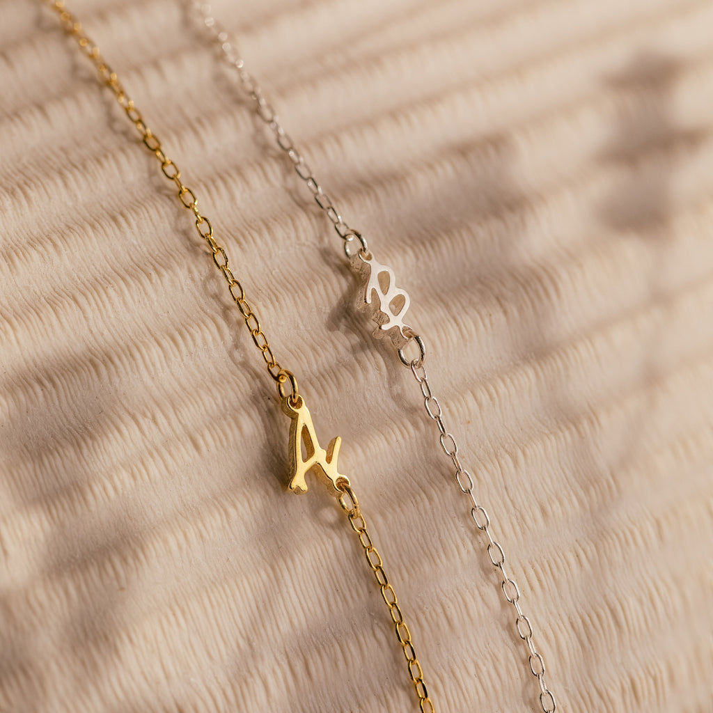 Gold and silver chain bracelets with letter A charms rest on a textured beige surface—perfect for stacking with your favorite initial jewelry or pairing with the Venice Sideways Initial Necklace.