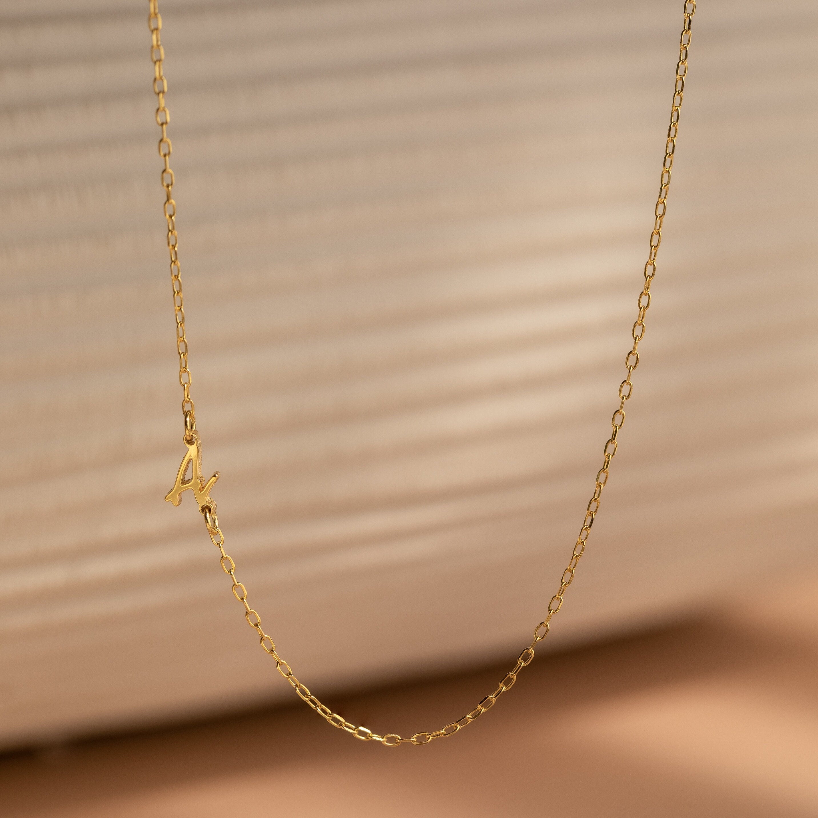 The Venice Sideways Initial Necklace features a delicate gold chain with a small sideways M pendant, perfect for lovers of subtle, personalized jewelry and beautifully showcased against a neutral background.