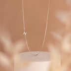 The Sideways Initial Necklace In Box Chain showcases a small letter H pendant, displayed elegantly on a soft beige background.