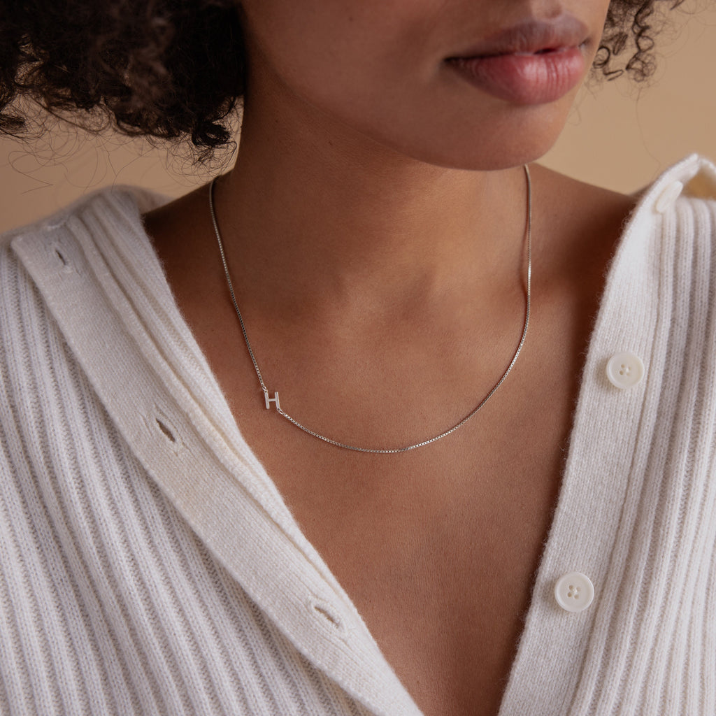 A woman wears a white sweater and the Sideways Initial Necklace In Box Chain, featuring a silver H pendant.