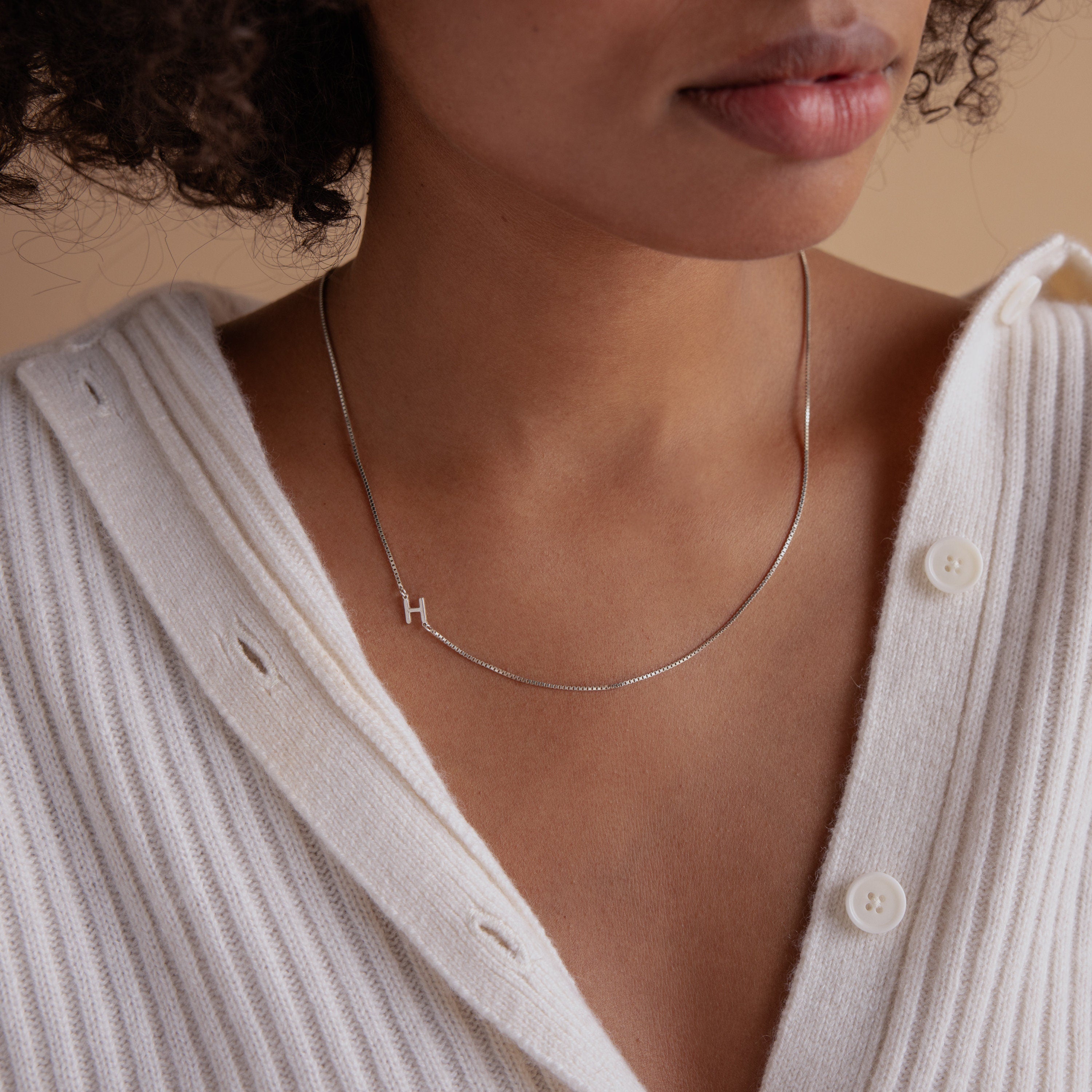 A woman wears a white sweater and the Sideways Initial Necklace In Box Chain, featuring a silver H pendant.